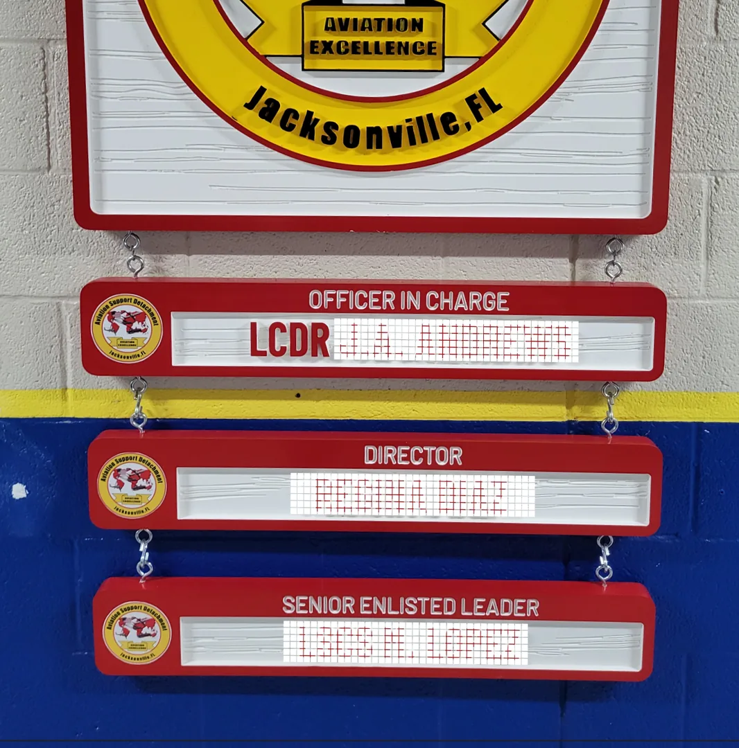 Unit crest above three red combined title and name plates reading Officer in Charge, Director and Senior Enlisted Leader, each hanging from the plate above